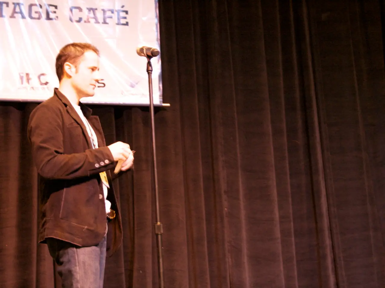 In this image there is a person standing , a mike with a mike stand, curtain, banner.