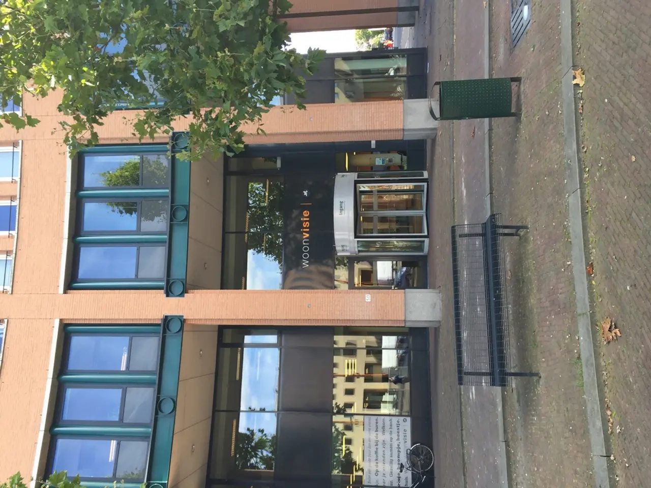 As we can see in the image there is a building, dustbin, bench, tree and a banner.