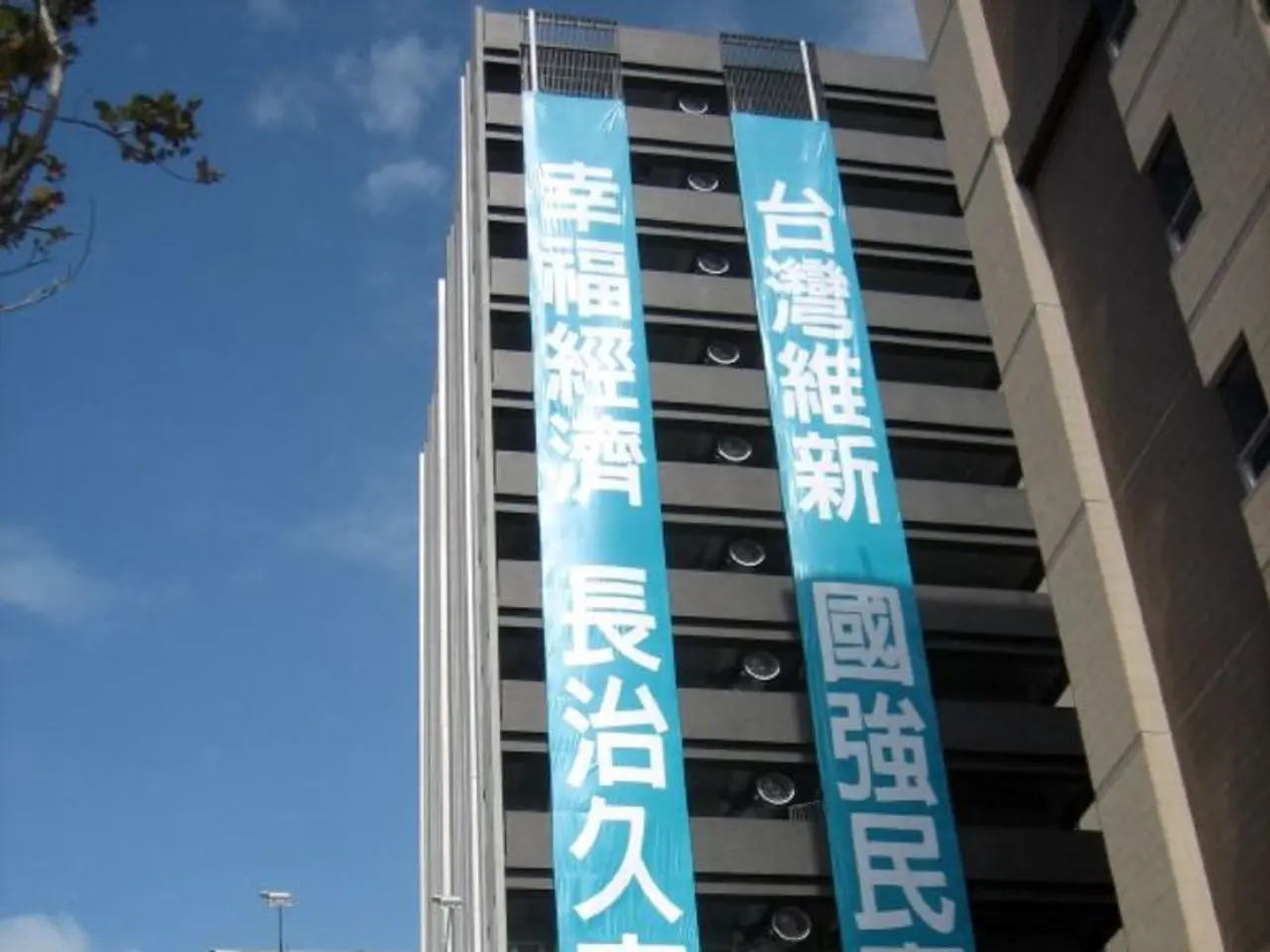 In this image there are buildings and we can see boards. In the background there are poles, trees...