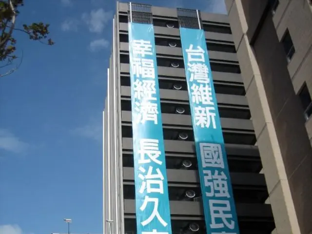 In this image there are buildings and we can see boards. In the background there are poles, trees...
